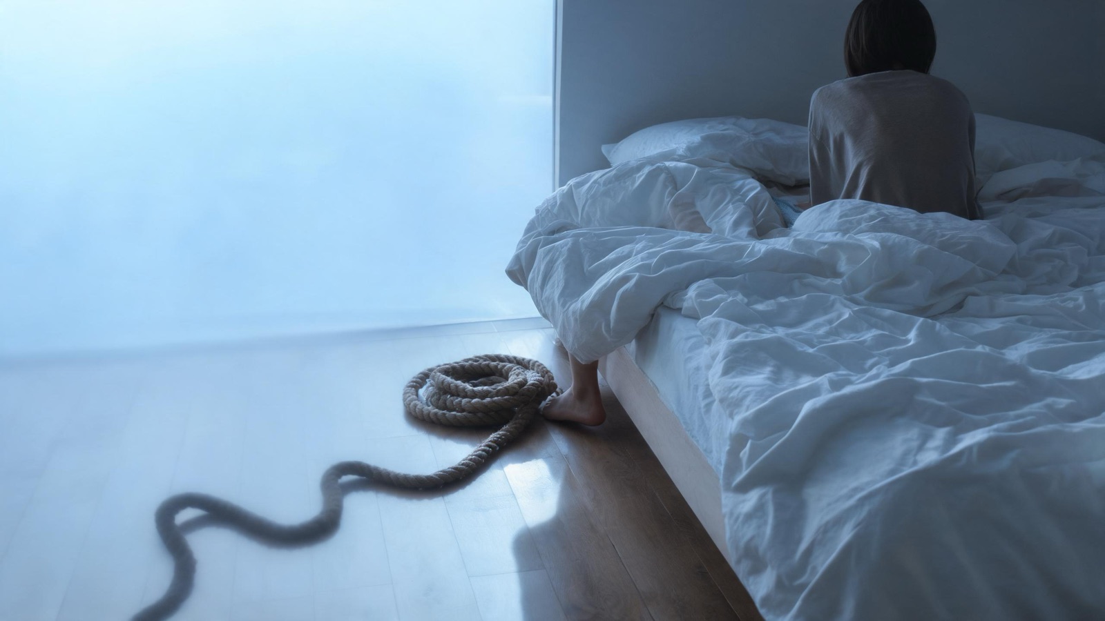 Un letto disfatto all'alba, con una corda arrotolata che proietta un'ombra sinuosa