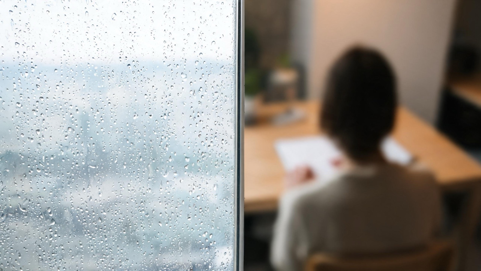 Gocce di pioggia sul vetro di una finestra, luce grigia e tepore sfocato all'interno