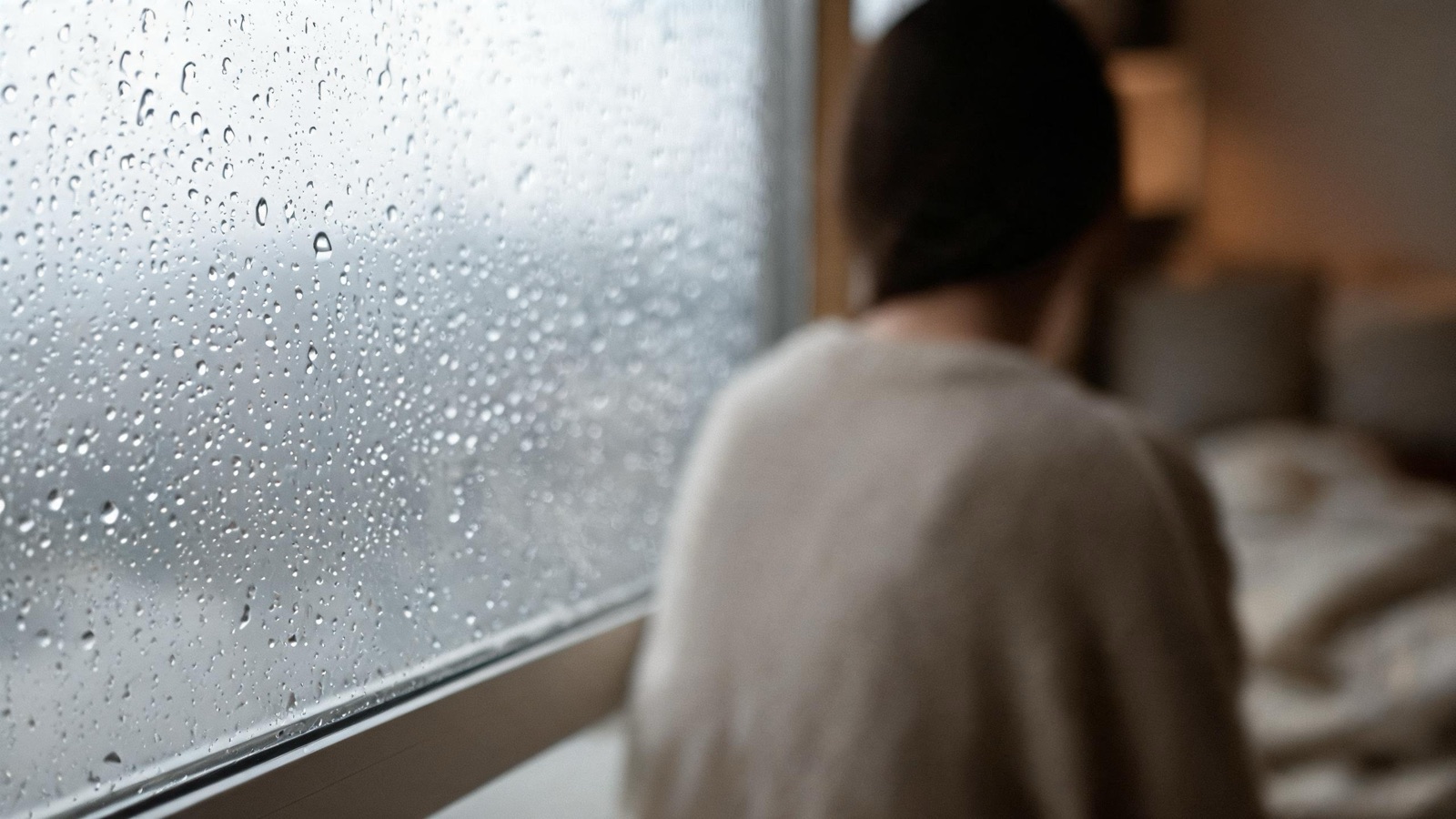 Gocce di pioggia sul vetro di una finestra, luce grigia e tepore sfocato all'interno
